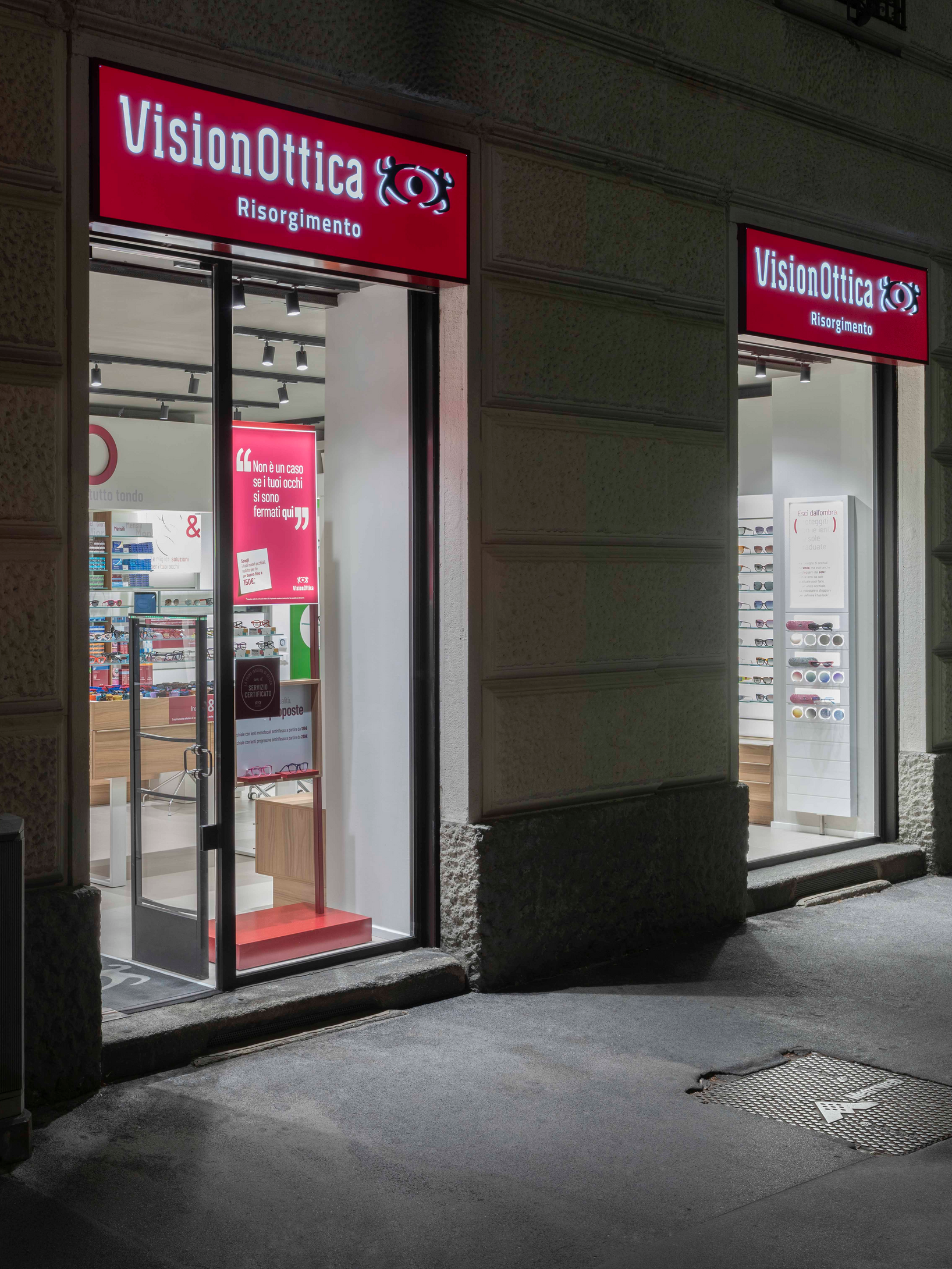 Facciata notturna del negozio VisionOttica Risorgimento di Milano su strada con due vetrine illuminate, insegna rossa e porta a vetri con pannello promozionale interno