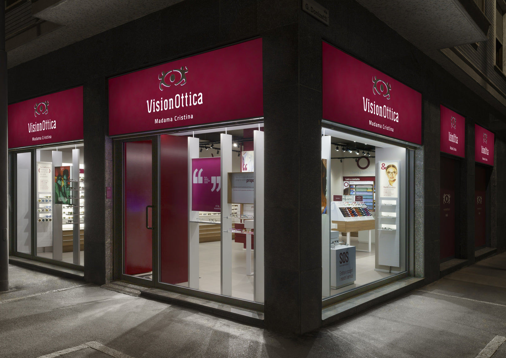 Facciata notturna del negozio VisionOttica Madama Cristina di Torino su strada con insegne bordeaux su tre lati, due vetrine illuminate e espositori di occhiali interni