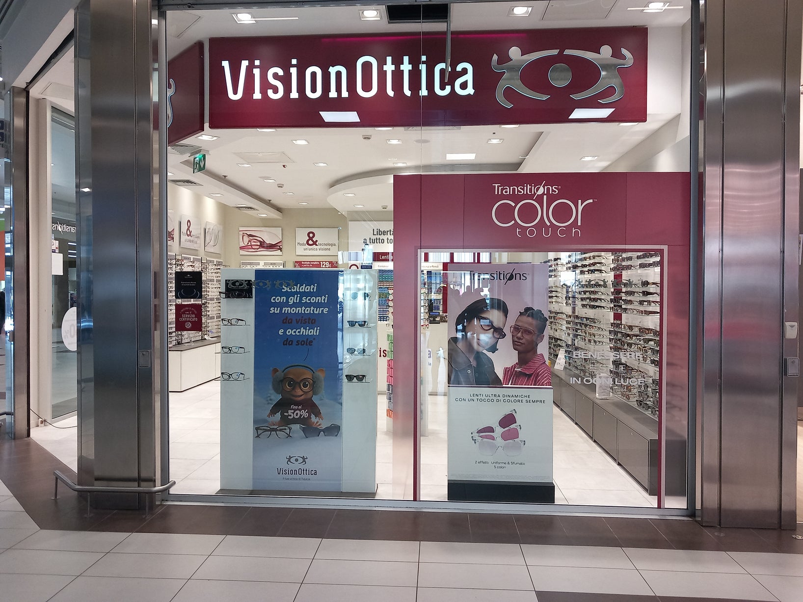 Ingresso del negozio VisionOttica Ossola di Crevoladossola in centro commerciale con insegna bordeaux, banner promozionale Transitions Color Touch e espositori di montature laterali