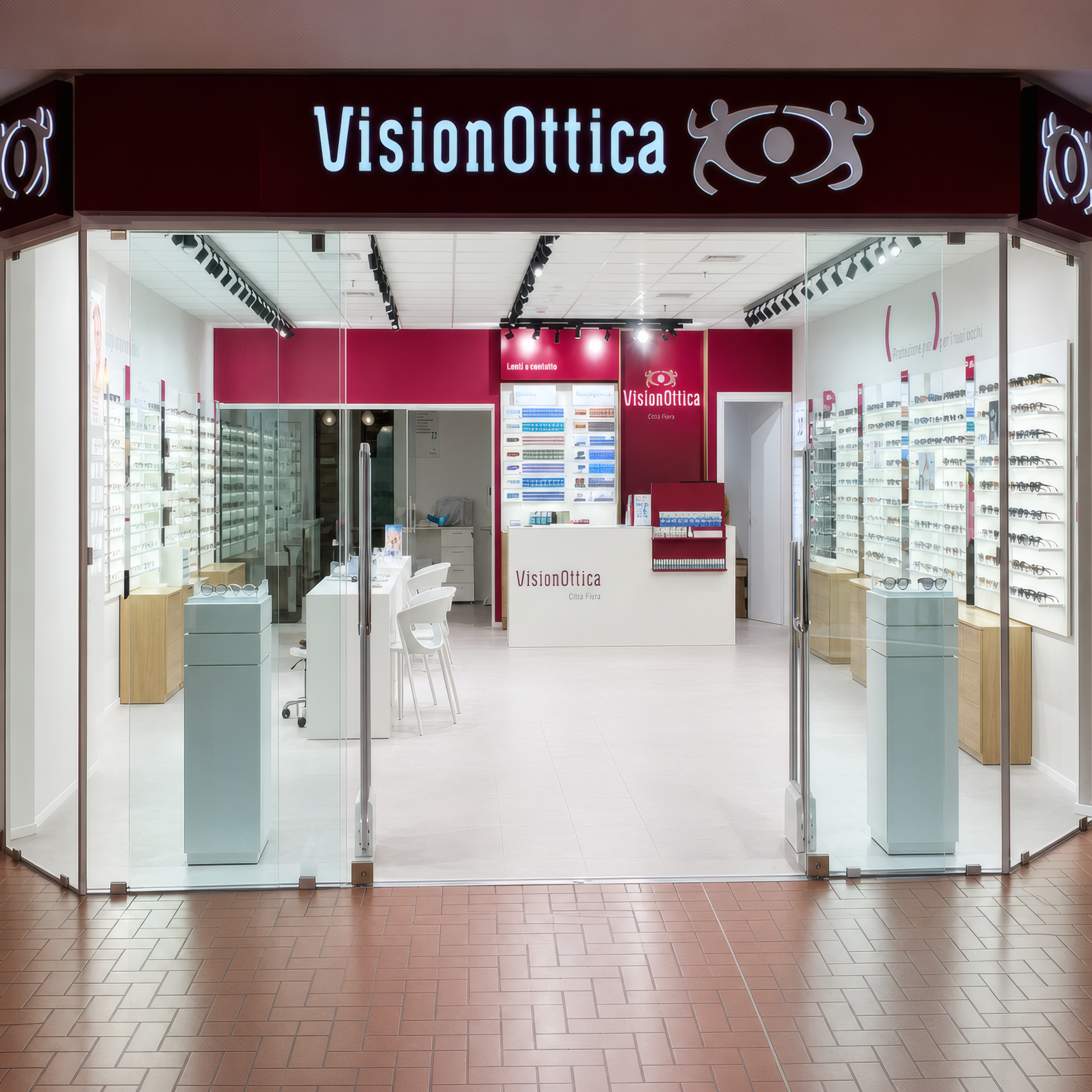 Ingresso del negozio VisionOttica Città Fiera di Udine in centro commerciale con insegna bianca su fondo bordeaux, interno bianco luminoso e espositori a parete