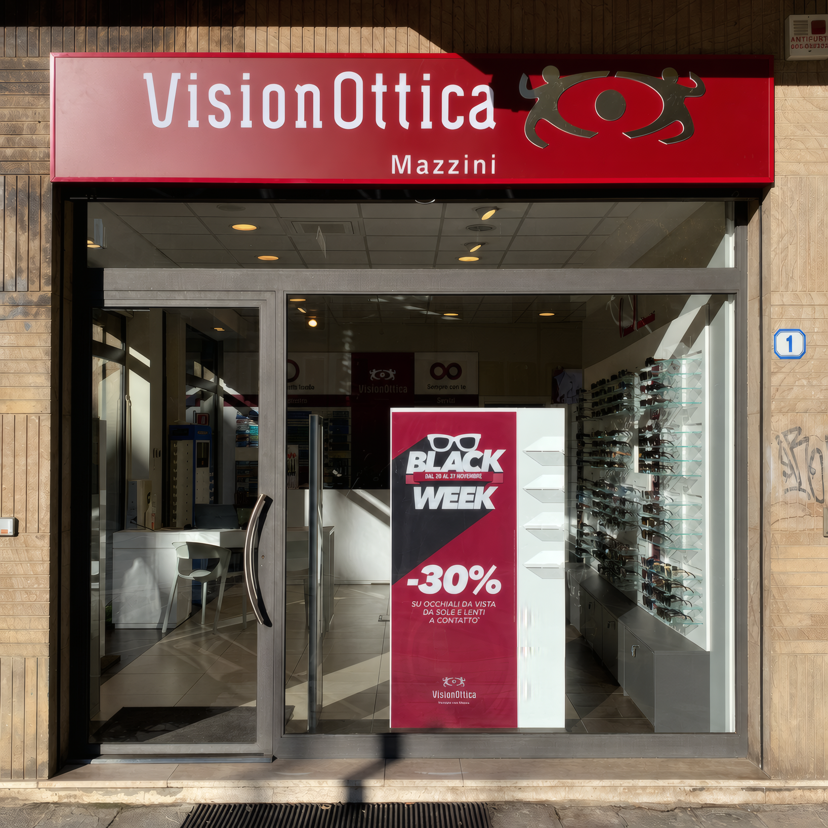 Facciata del negozio VisionOttica Mazzini di Padova su strada con insegna bordeaux, porta a vetri centrale e vetrina con banner Black Week -30% e espositori di occhiali
