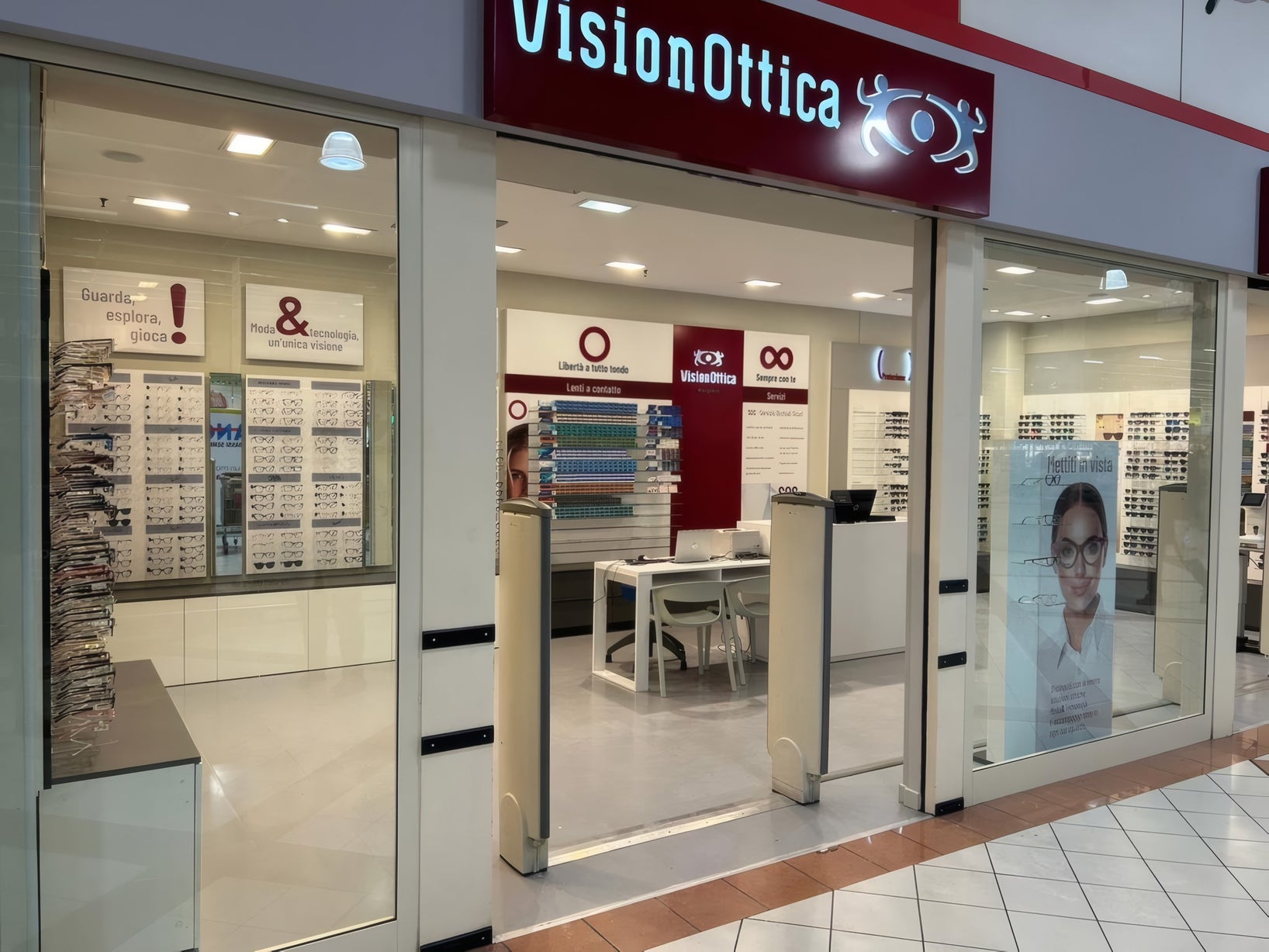 Ingresso del negozio VisionOttica Marghera di Marghera in centro commerciale con insegna bordeaux, doppia apertura a vetri, scaffali di occhiali a parete e poster pubblicitario con optometrista