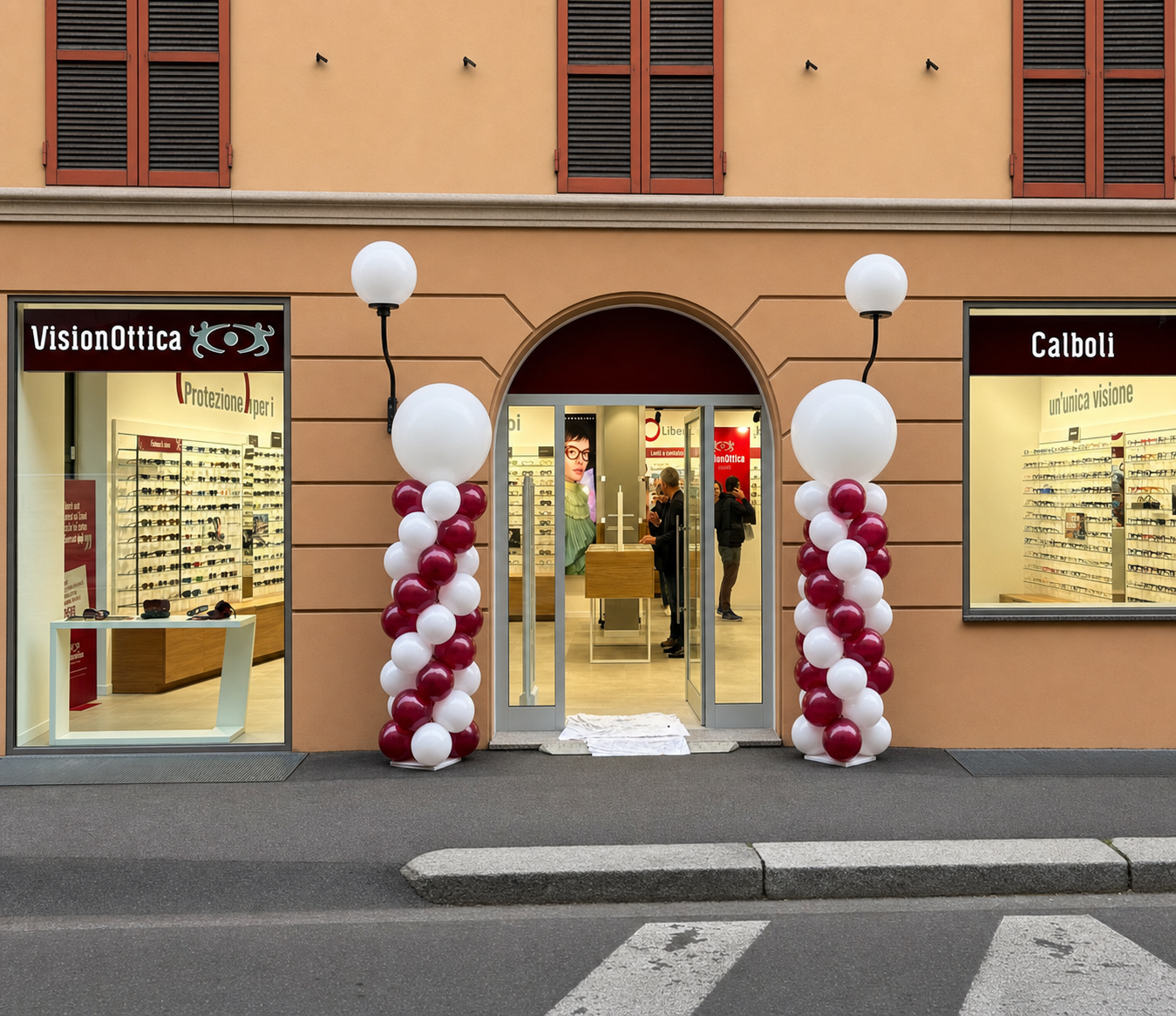 Ingresso del negozio VisionOttica Calboli di Forlì in centro storico su strada con facciata color ocra, portale ad arco, insegna bordeaux e colonne decorate con palloncini bordeaux e bianchi per inaugurazione