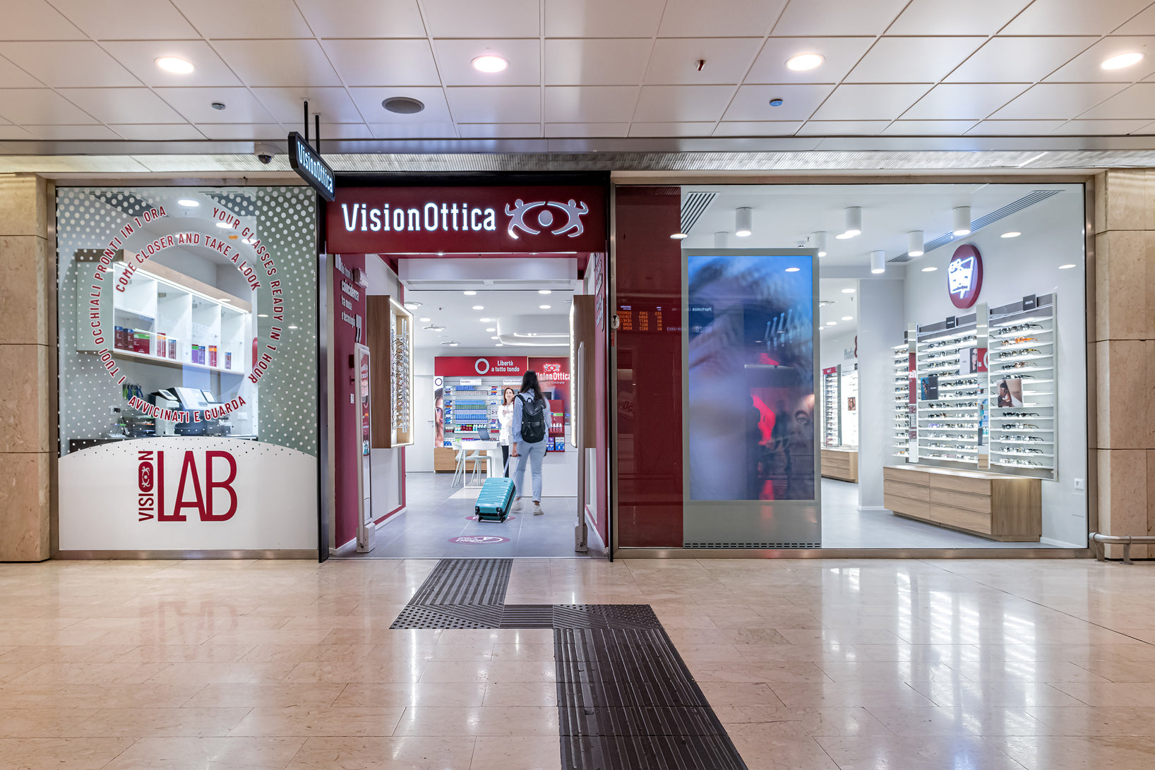 Ingresso del negozio VisionOttica Milano Centrale in centro commerciale alla Stazione Centrale di Milano con insegna bordeaux, totem VisionLab a sinistra, schermo digitale centrale e scaffali di occhiali