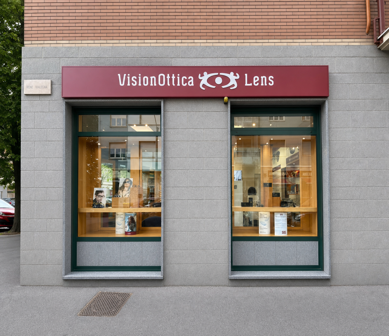 Facciata del negozio VisionOttica Lens di Torino su strada con insegna bordeaux orizzontale, due vetrine rettangolari con espositori di montature e rivestimento in pietra grigia