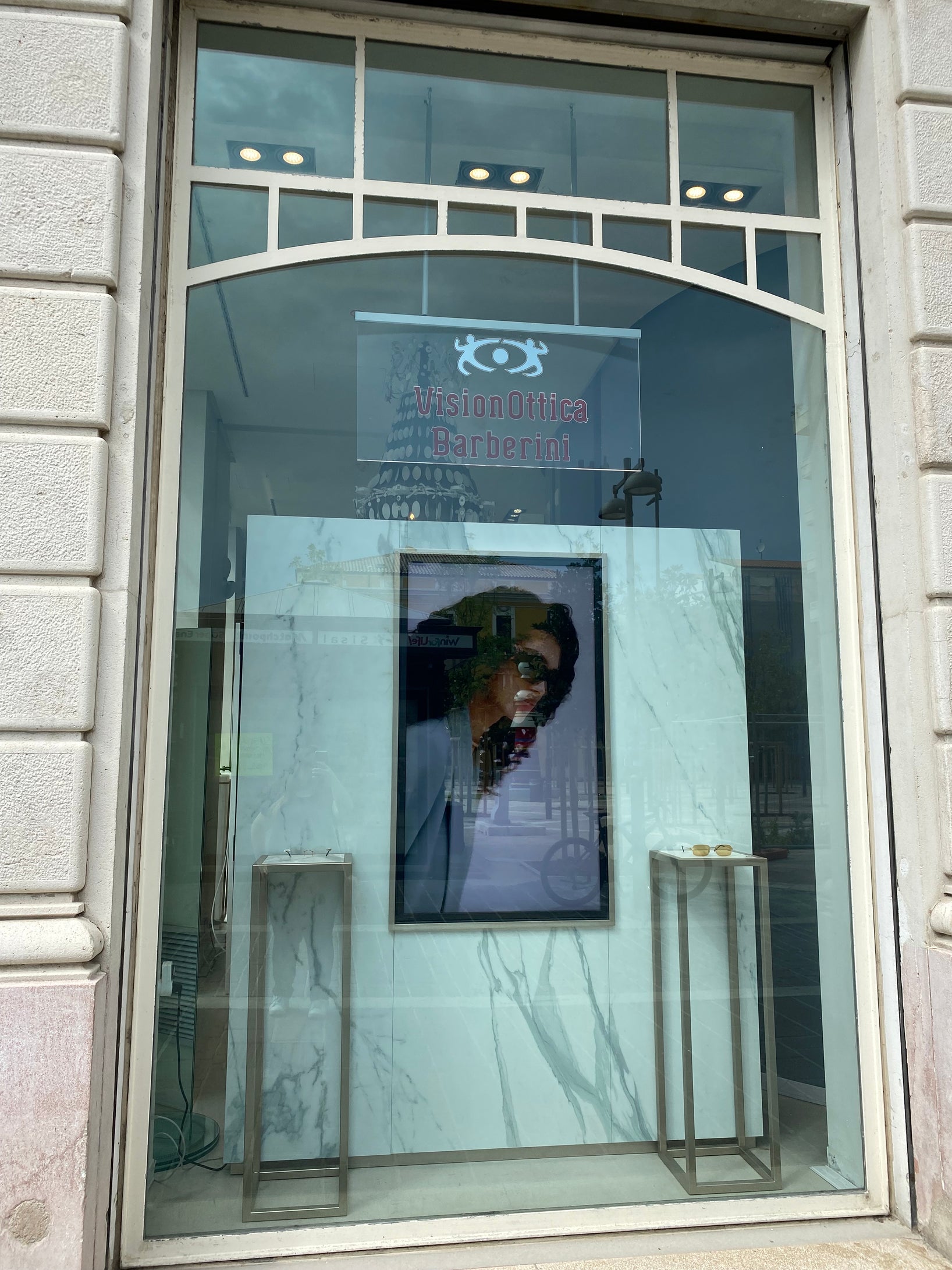 Vetrina del negozio VisionOttica Barberini di Pescara su strada con design minimal in marmo bianco, logo inciso sul vetro, schermo digitale centrale e scaffali metallici laterali vuoti