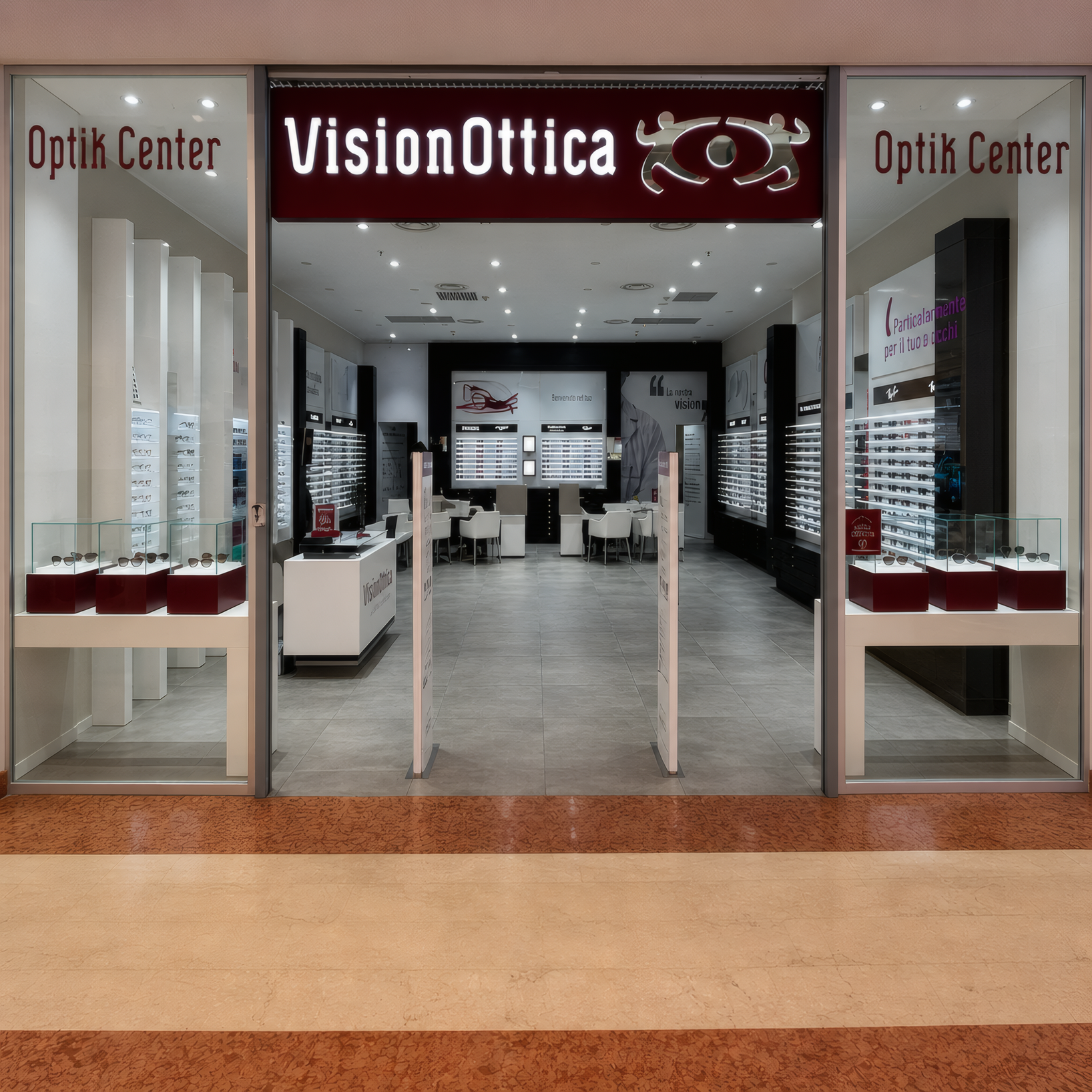 Ingresso del negozio VisionOttica Optik Center di Varese in centro commerciale con insegna bordeaux, porta a vetri centrale e bancone con espositori di occhiali disposti a semicerchio