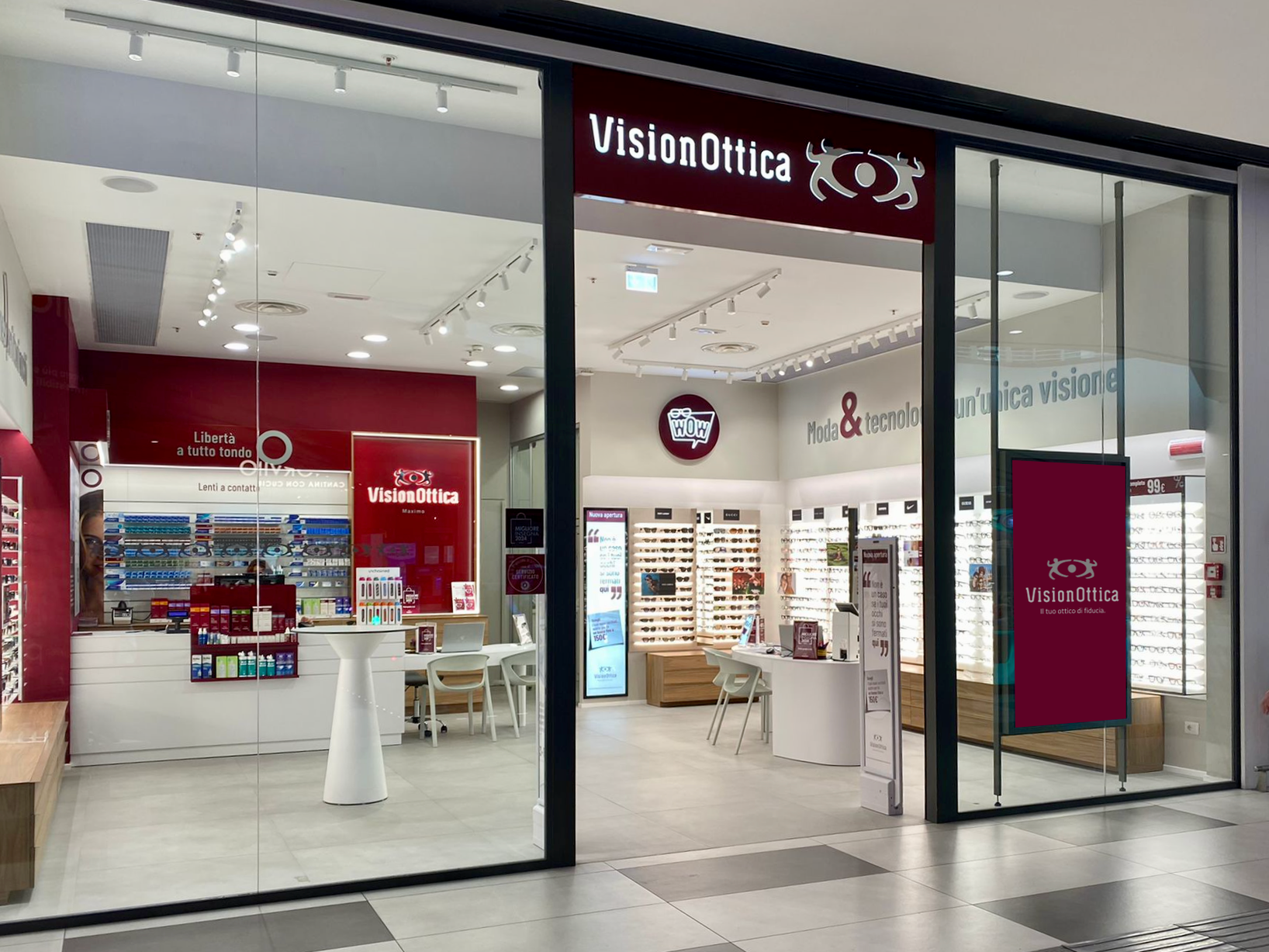Ingresso del negozio VisionOttica Maximo di Roma in centro commerciale con insegna bordeaux, ampia apertura a vetrate, bancone centrale rosso e scaffali di montature sulle pareti laterali