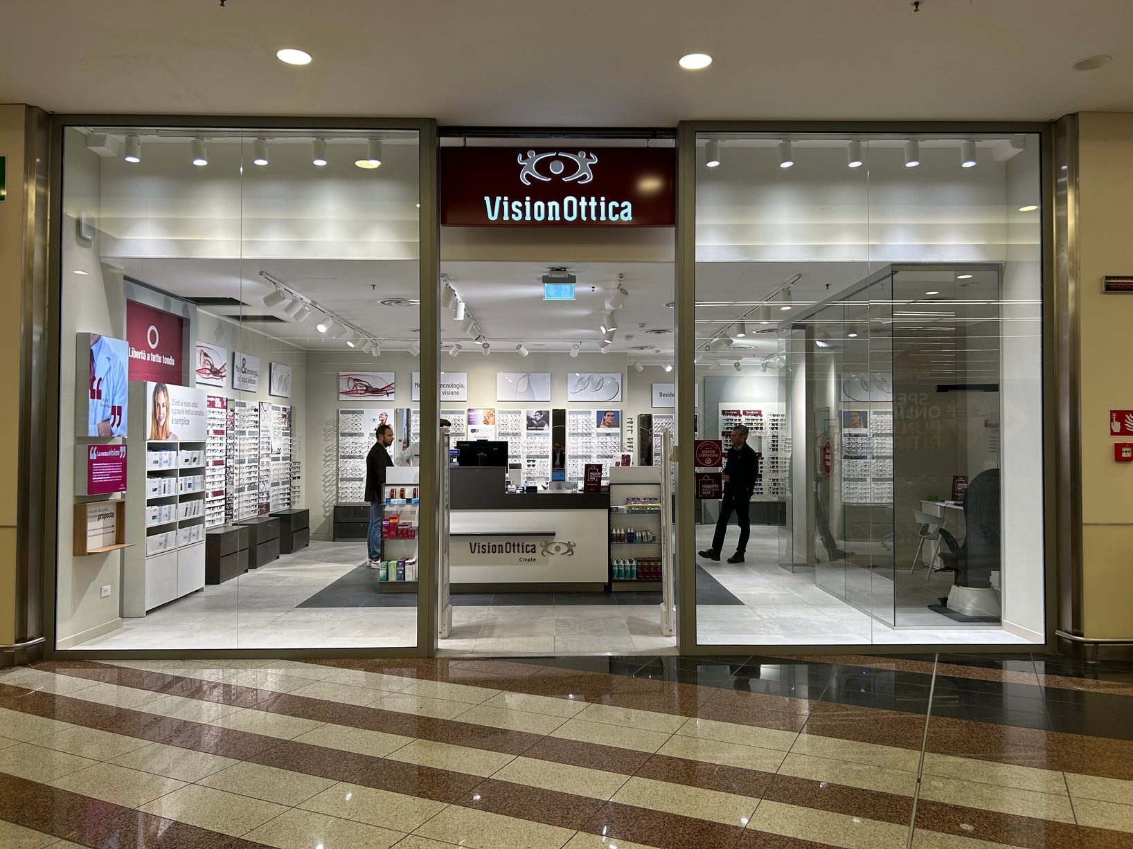 Ingresso del negozio VisionOttica Civate di Civate in centro commerciale con insegna bordeaux su fondo bianco, doppia vetrata e interno con espositori di occhiali e personale al bancone