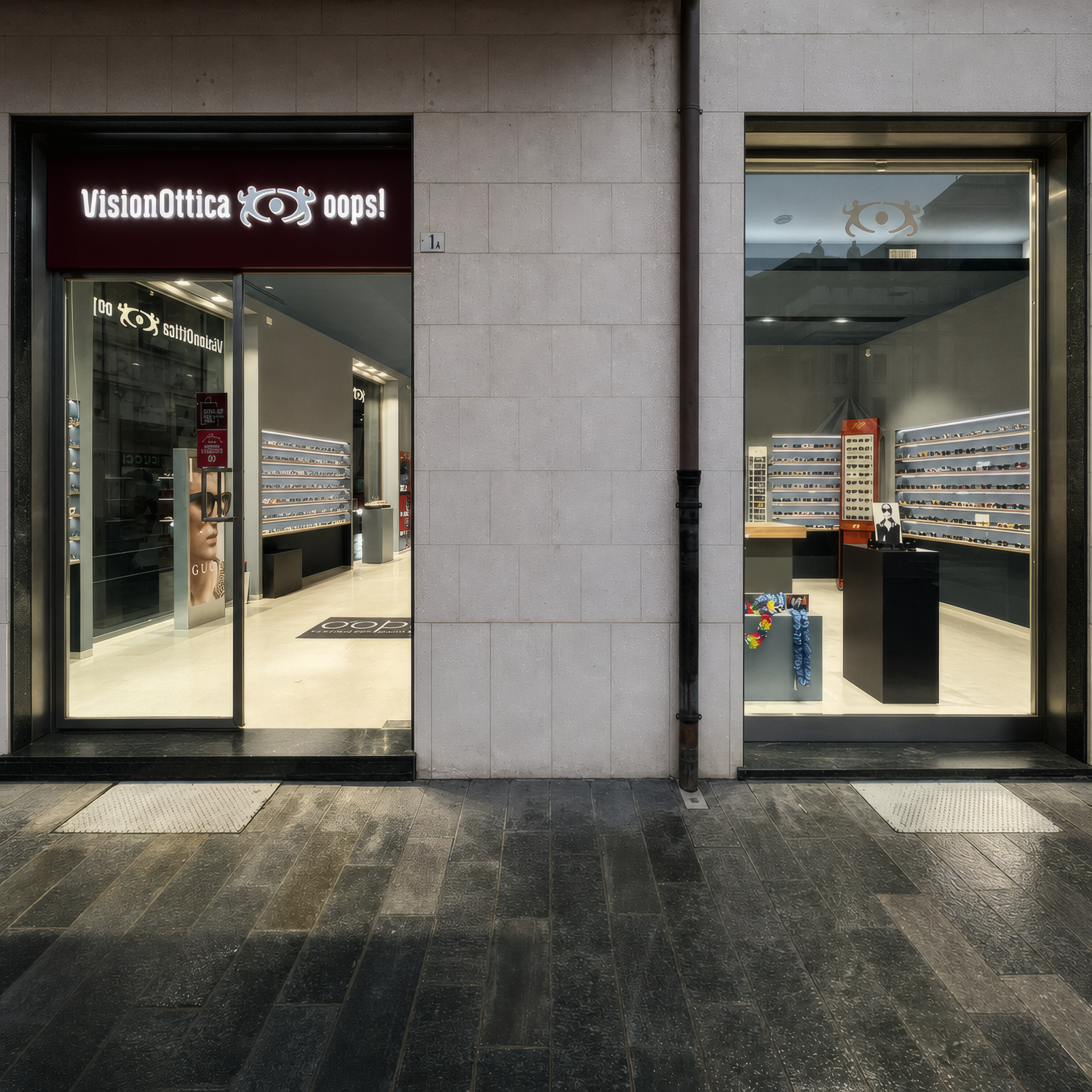 Facciata del negozio VisionOttica Oops! di Alba su via pedonale con rivestimento in pietra scura, doppia vetrina illuminata con espositori di montature e insegna rossa con nome del negozio