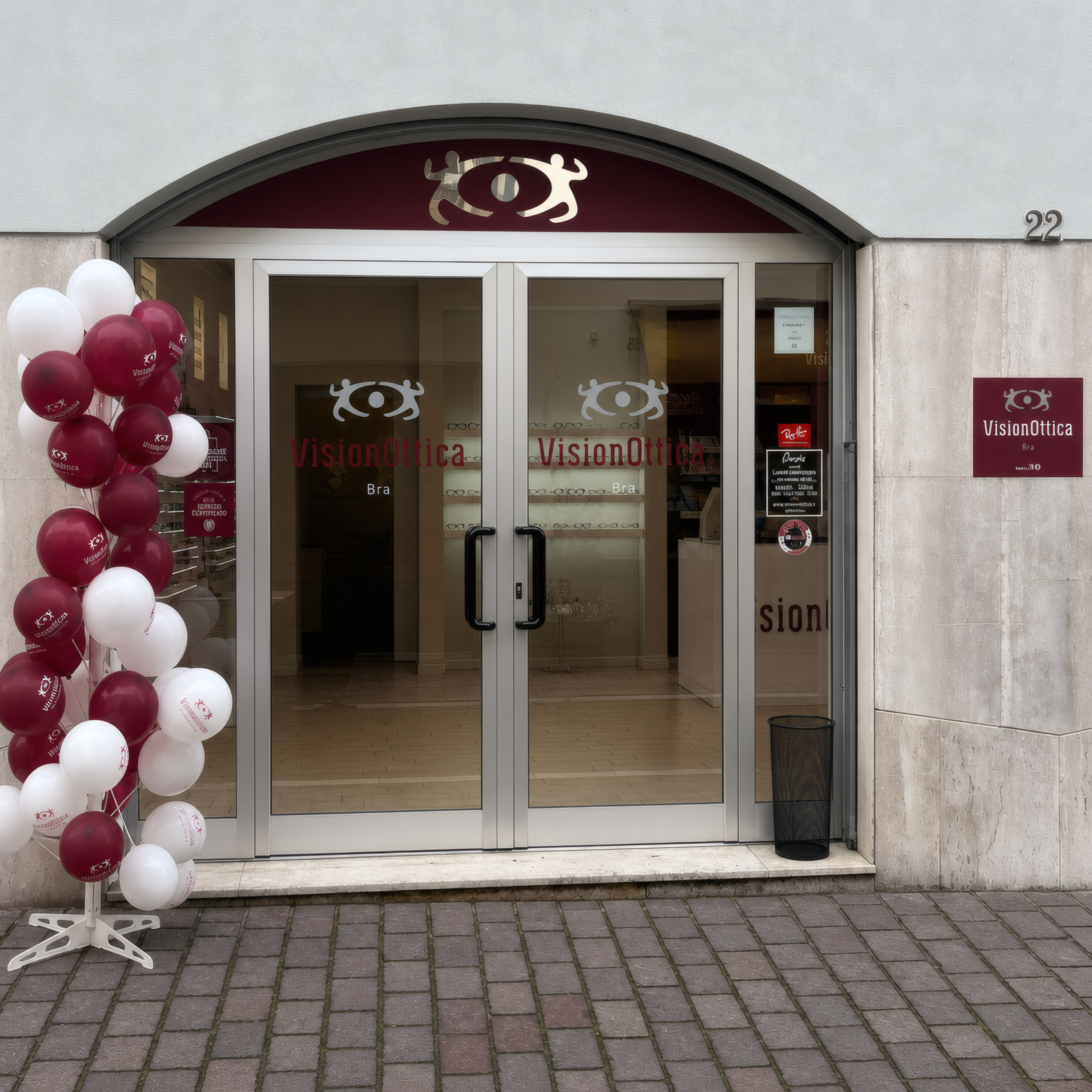 Ingresso del negozio VisionOttica Bra di Bra su via pedonale con portale ad arco in pietra chiara, insegna bordeaux, porta a vetri con decorazioni di palloncini per inaugurazione