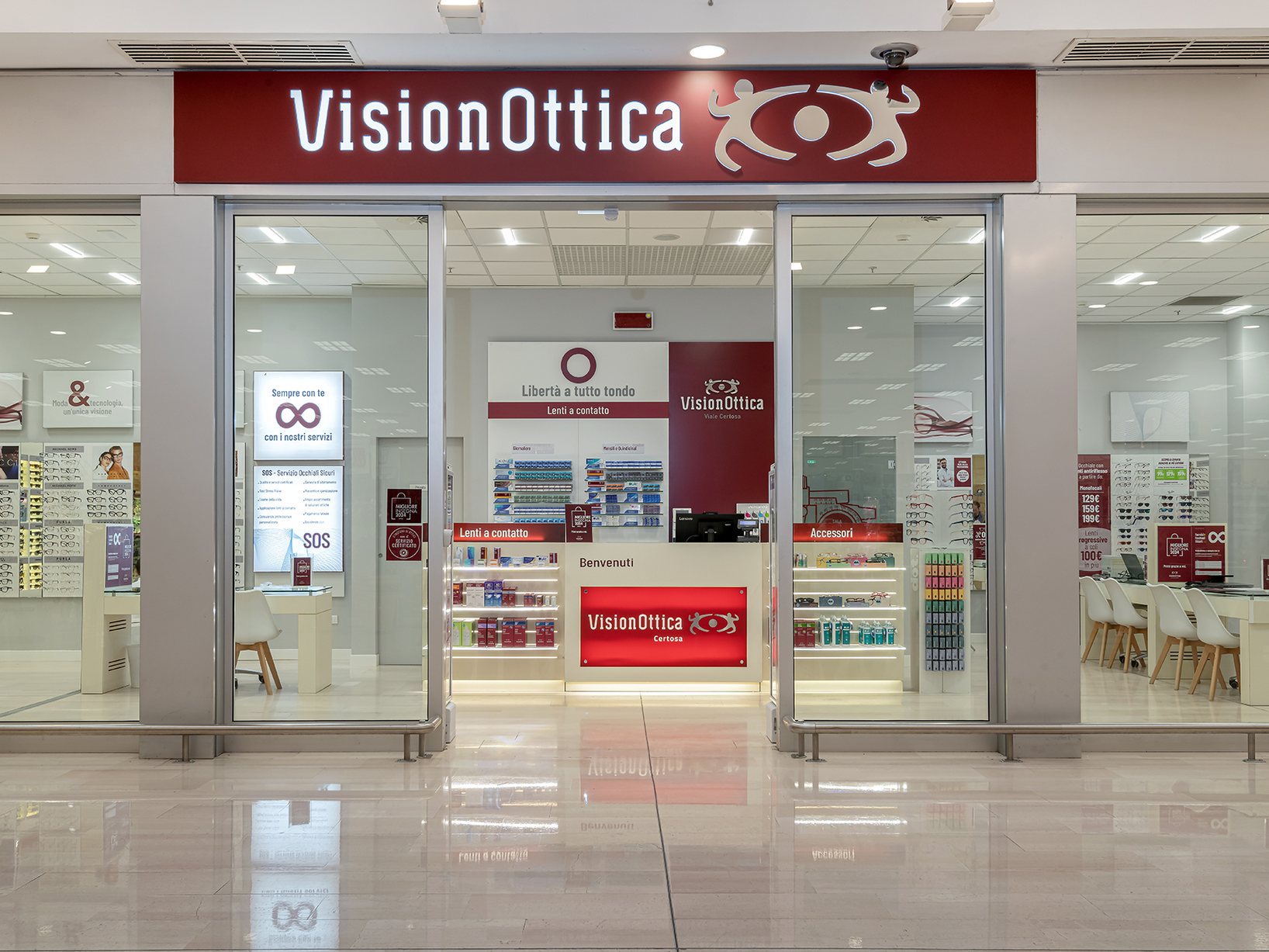 Ingresso del negozio VisionOttica Certosa di Milano in centro commerciale con ampia insegna bordeaux, vetrata a tutta altezza, bancone di accoglienza centrale e espositori di montature e lenti a contatto