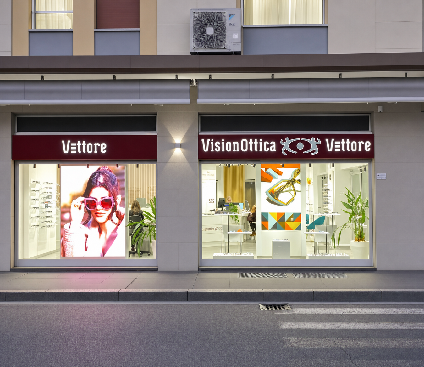Facciata del negozio VisionOttica Vettore di Varese su strada con doppia vetrina illuminata, insegna bordeaux e poster pubblicitario con modella