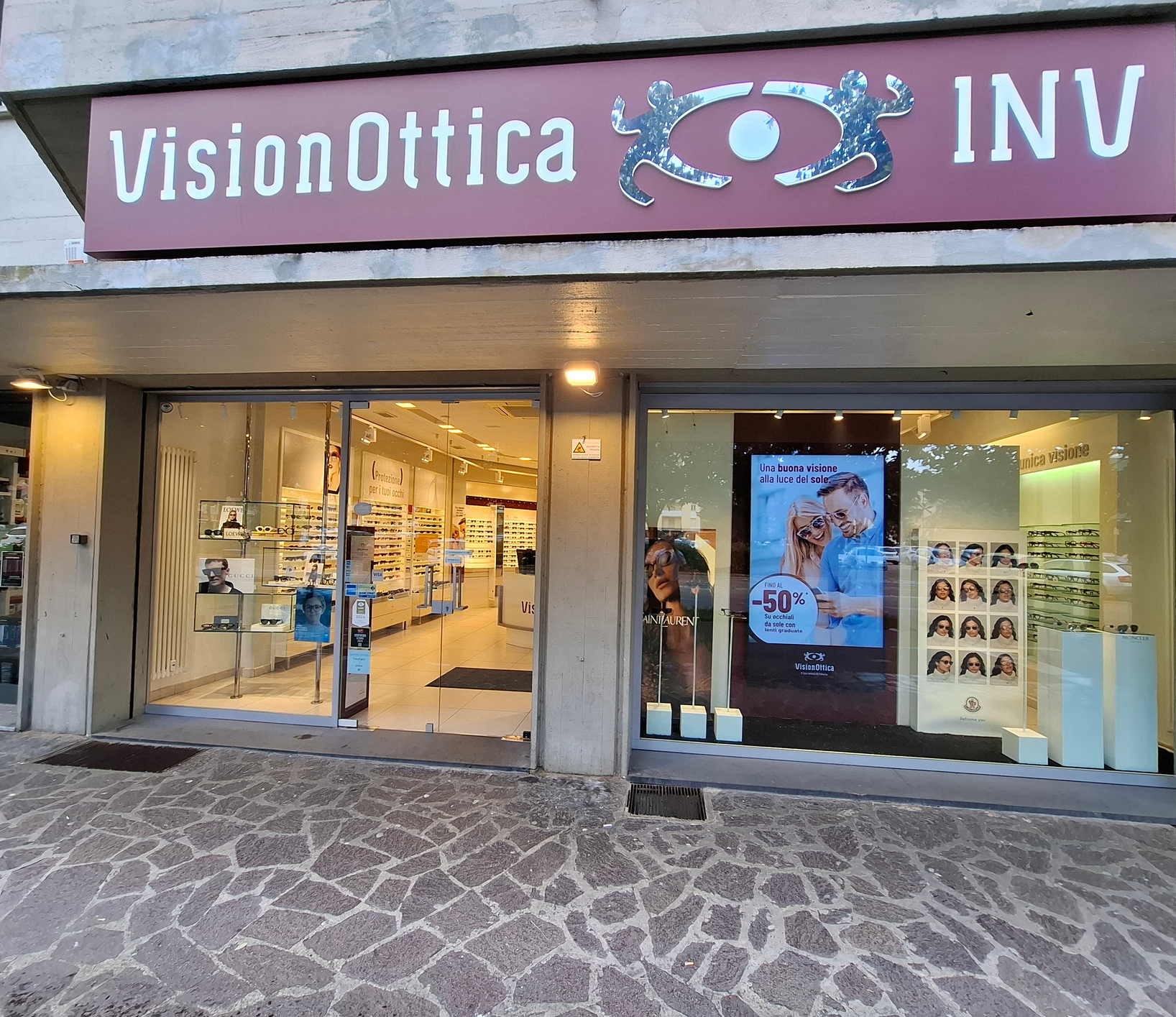 Facciata del negozio VisionOttica INV di Scandicci su via pedonale con insegna bordeaux ampia, doppia vetrina e banner promozionale con sconto -50%