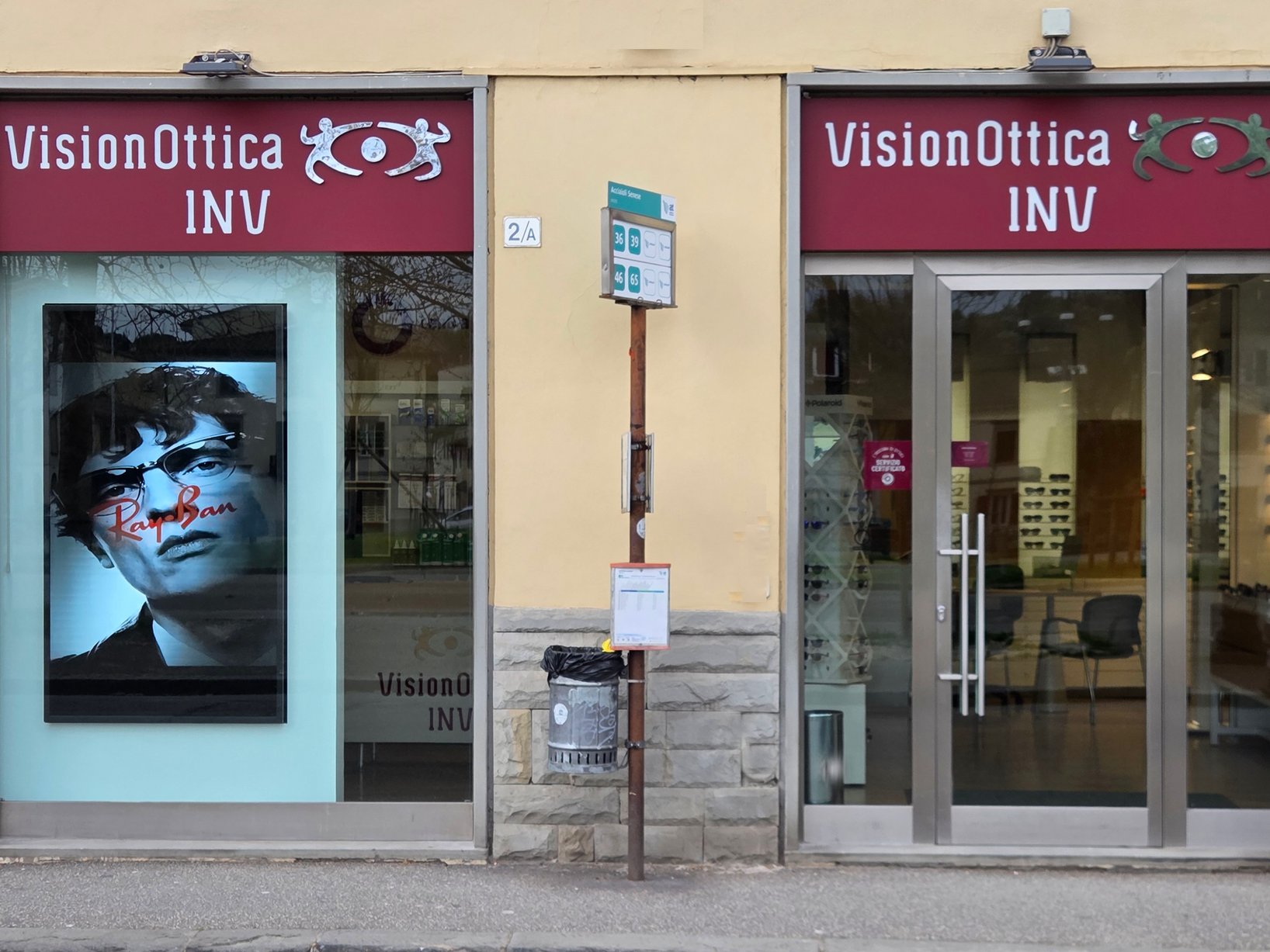 Facciata del negozio VisionOttica INV di Firenze su strada con insegna bordeaux larga, vetrina con poster Ray-Ban e porta a vetri