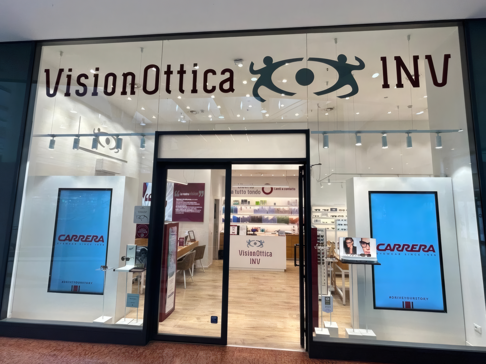 Ingresso del negozio VisionOttica INV di Firenze in centro commerciale con vetrina a tutta altezza, insegna bianca e schermi digitali Carrera laterali