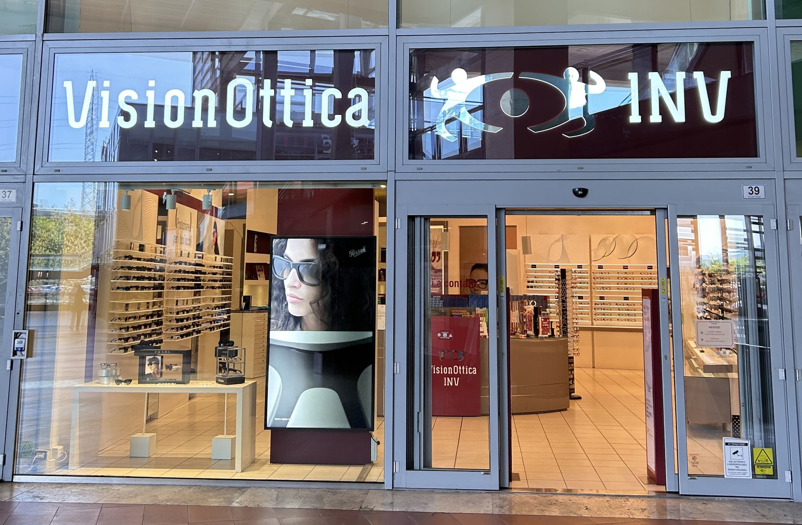 Ingresso del negozio VisionOttica INV di Prato in centro commerciale con vetrina panoramica, insegna luminosa e poster di donna con occhiali da sole