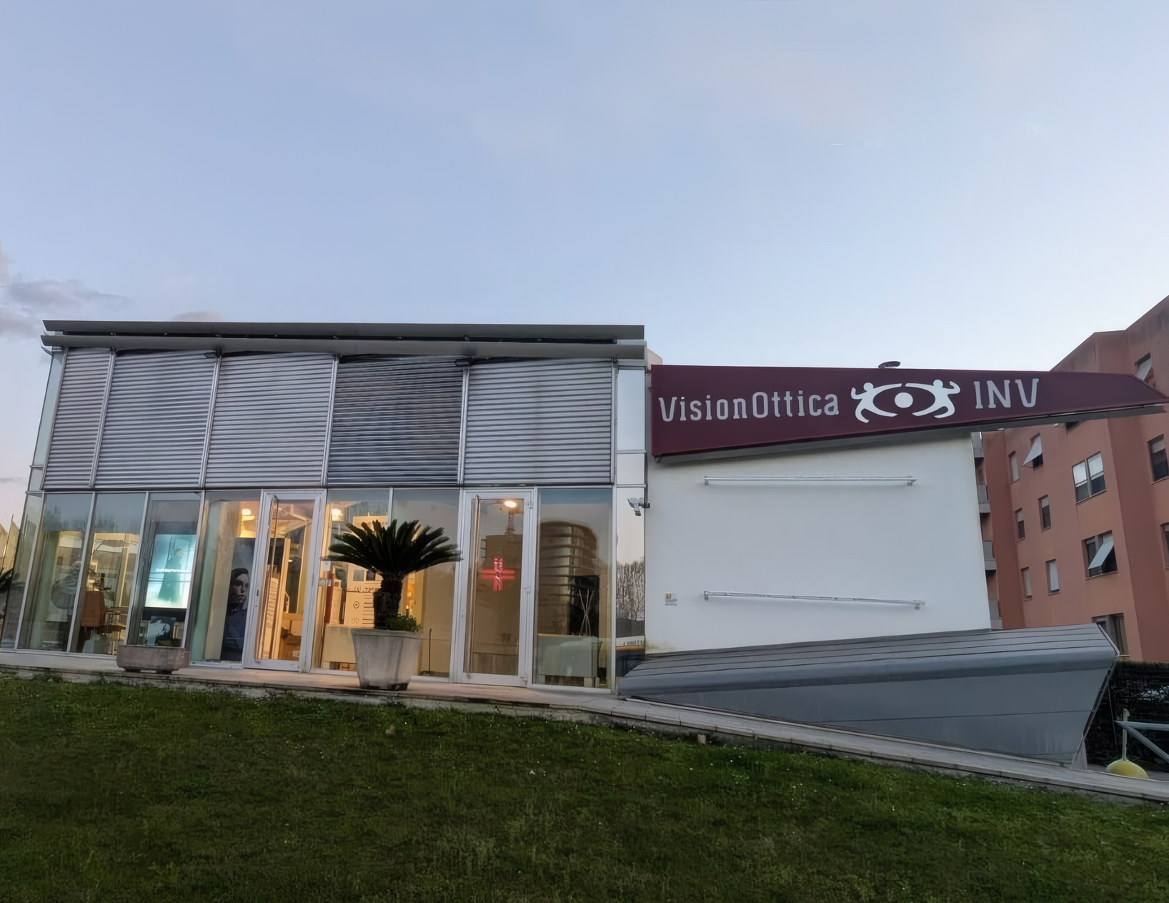 Esterno del negozio VisionOttica INV di Pisa su strada con ampia vetrata, insegna bianca su tetto ribassato grigio e parcheggio antistante