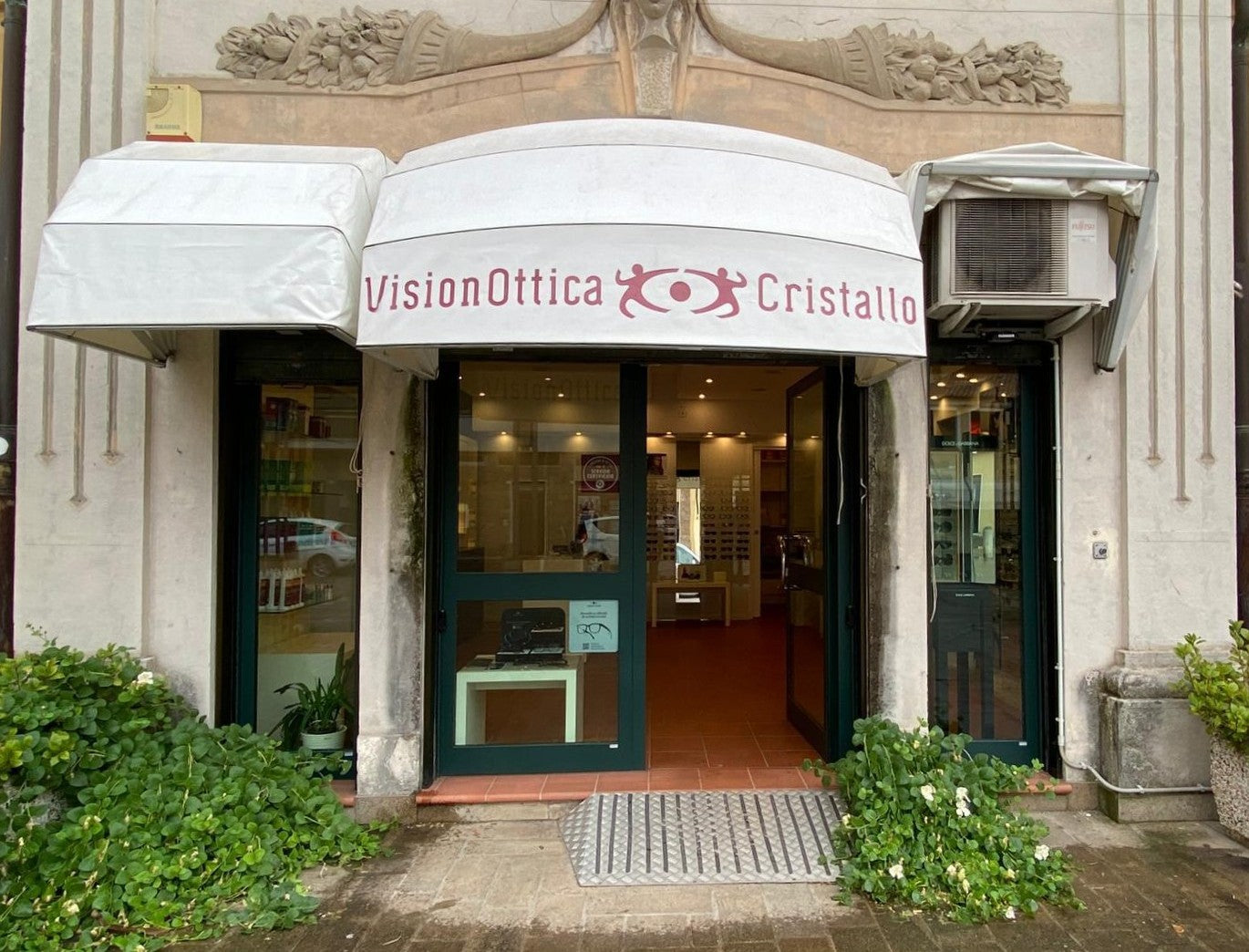 Ingresso del negozio VisionOttica Cristallo di Monselice in centro storico su edificio liberty con tenda avvolgente bianca, porta verde e vegetazione all'ingresso