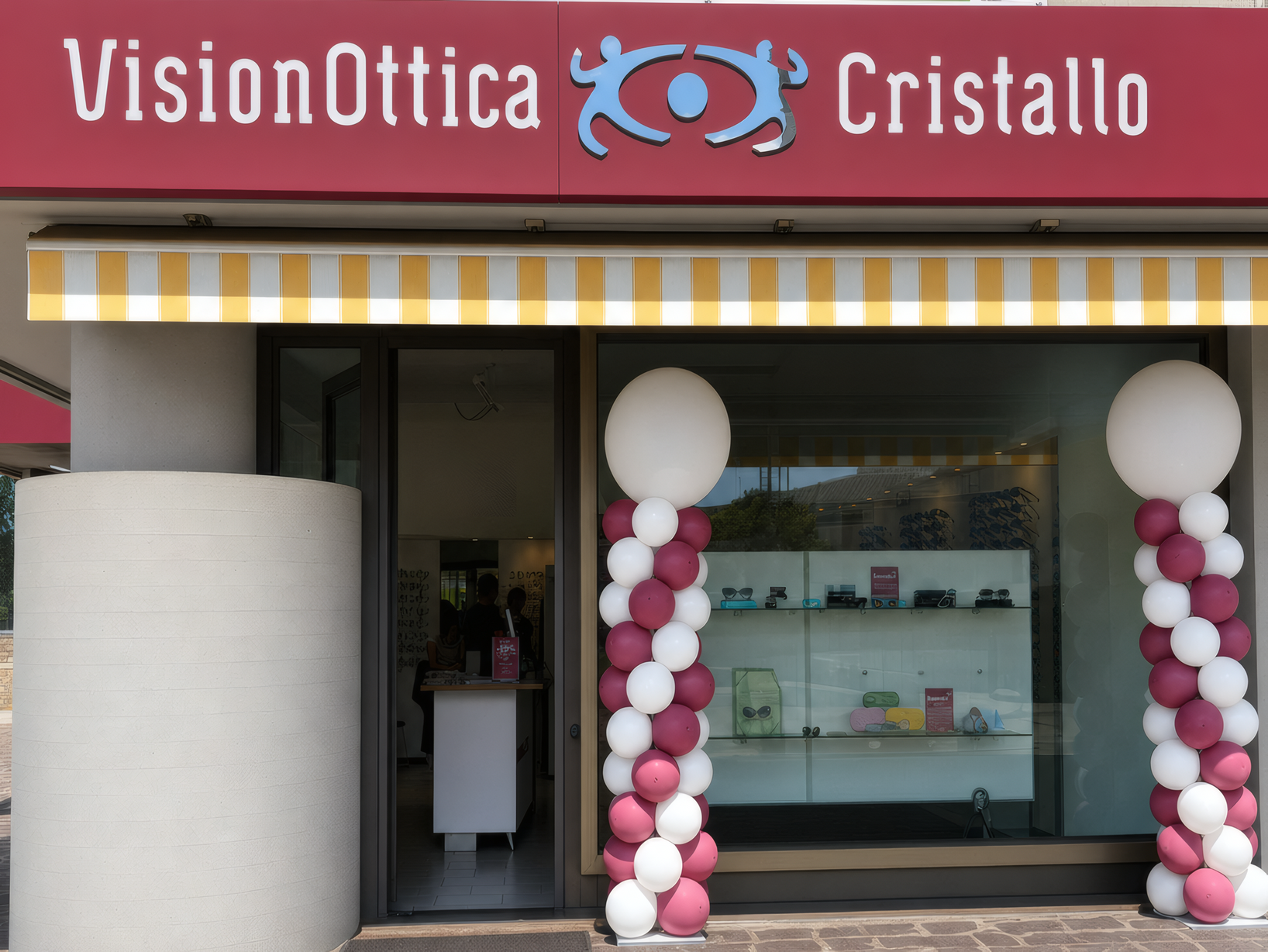 Facciata del negozio VisionOttica Cristallo di Conselve su strada con tenda a righe gialle e bianche, insegna bordeaux e palloncini decorativi all'inaugurazione