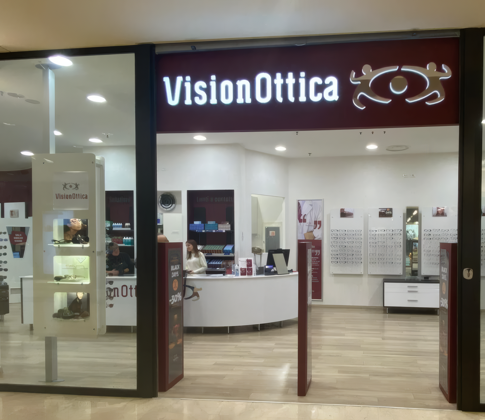 Ingresso del negozio VisionOttica Sacile di Sacile in centro commerciale con vetrina ampia, insegna bianca su sfondo scuro e espositori di montature