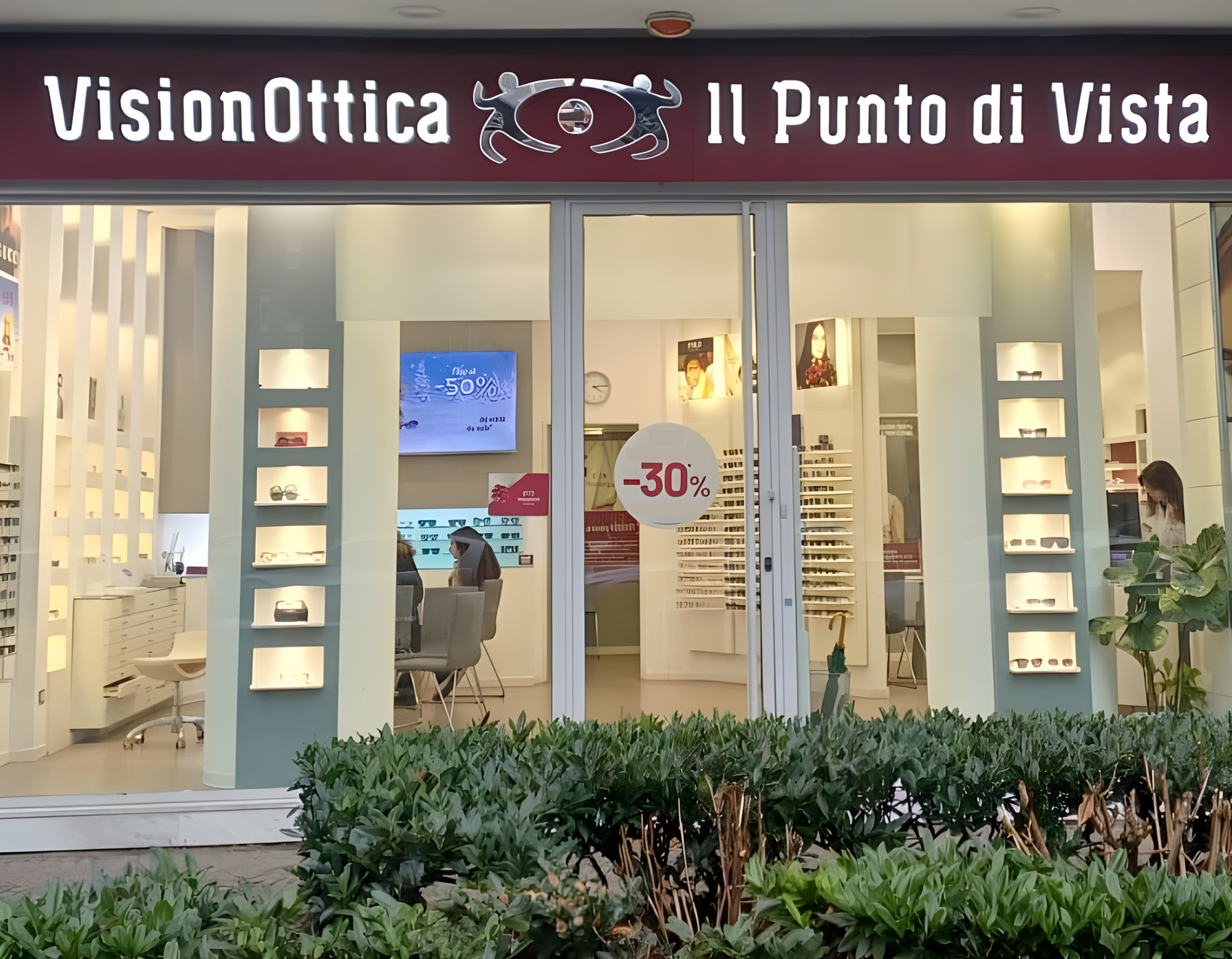 Vetrina del negozio VisionOttica Il Punto di Vista di Milano su strada con insegna bordeaux, espositori luminosi bianchi laterali, banner sconto -30% e display digitale interno