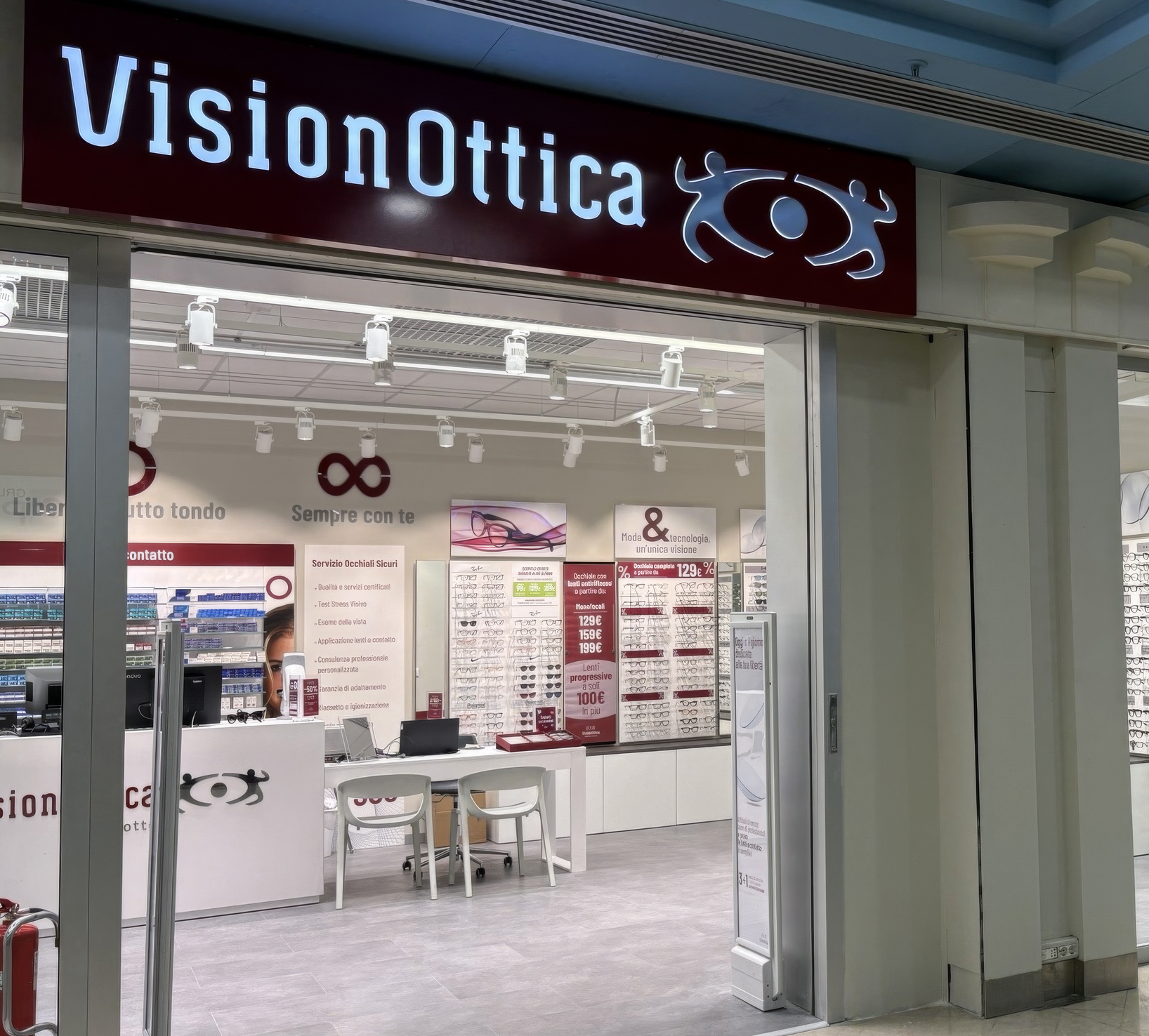 Ingresso del negozio VisionOttica Giotto di Padova in centro commerciale con grande insegna a LED bordeaux, bancone centrale bianco, roll-up promozionali e parete espositori di montature