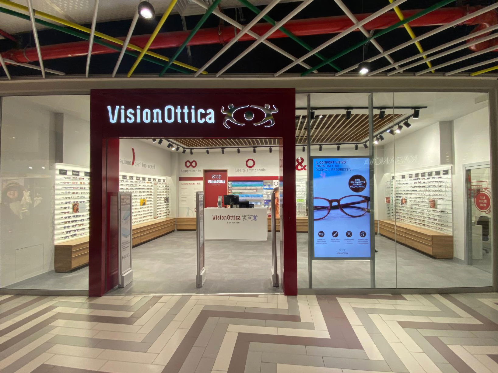 Ingresso del negozio VisionOttica Fanocenter di Fano in centro commerciale con insegna bordeaux illuminata, ampie vetrate trasparenti, espositori lineari di montature e display digitale laterale