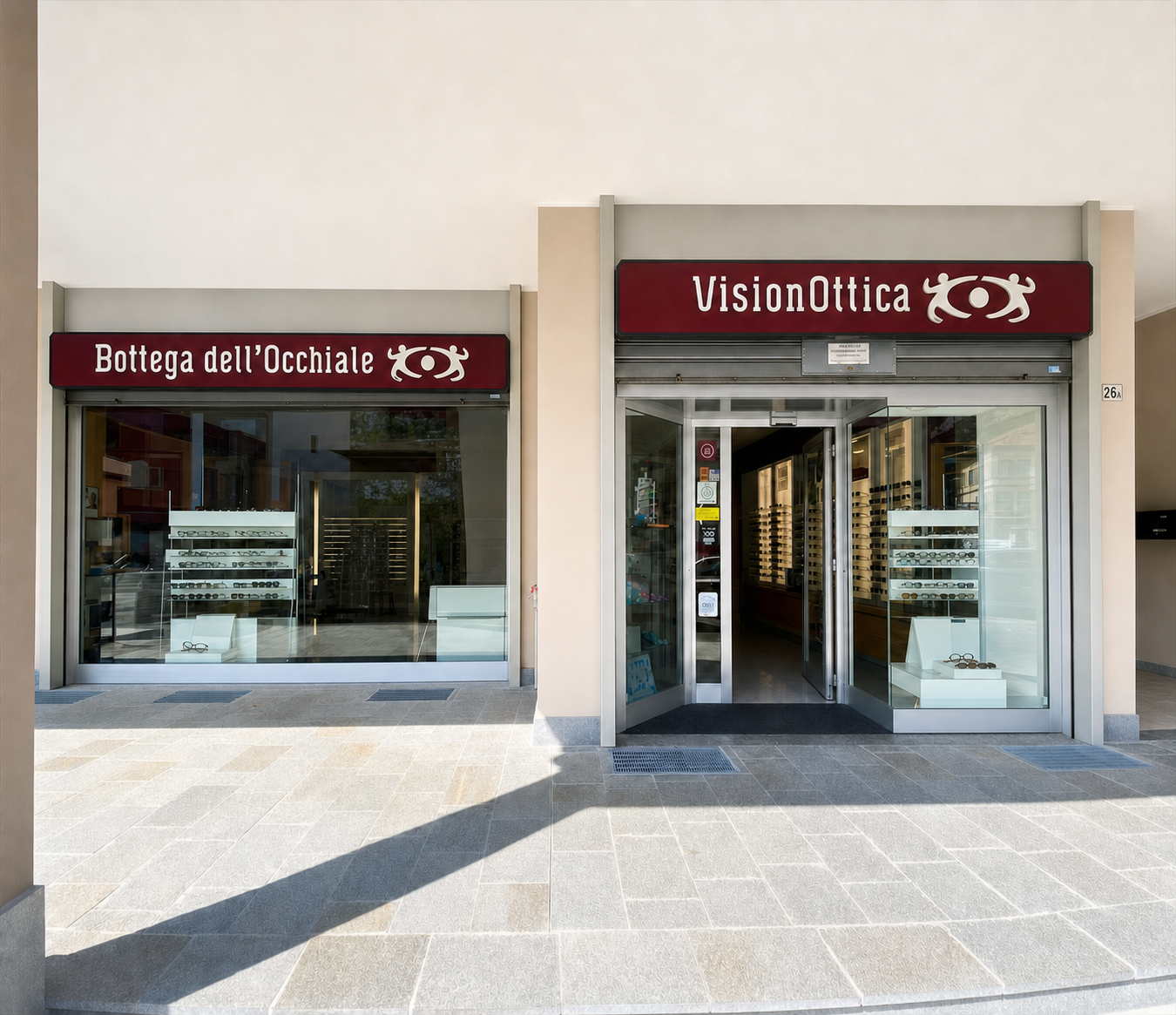 Facciata del negozio VisionOttica Bottega dell'Occhiale di Calolziocorte sotto porticato con doppia insegna bordeaux, porta automatica a vetri centrale e vetrine laterali con espositori di montature