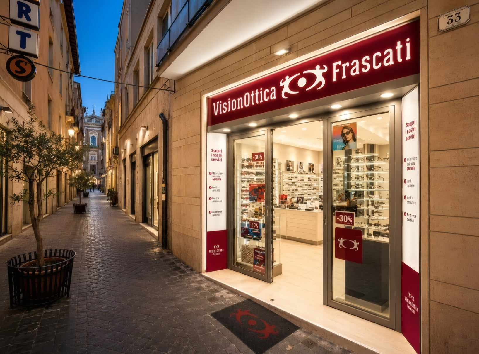 Facciata del negozio VisionOttica Frascati in centro storico su via pedonale lastricata al crepuscolo con insegna bordeaux illuminata, vetrina con espositori e banner sconto -30%