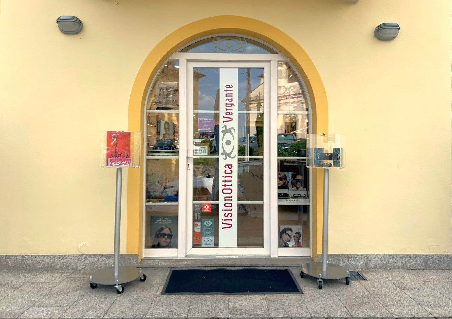 Ingresso del negozio VisionOttica Vergante di Nebbiuno su strada in centro con portale ad arco giallo, porta a vetri con insegna verticale, due espositori da terra con brochure e vetrine laterali