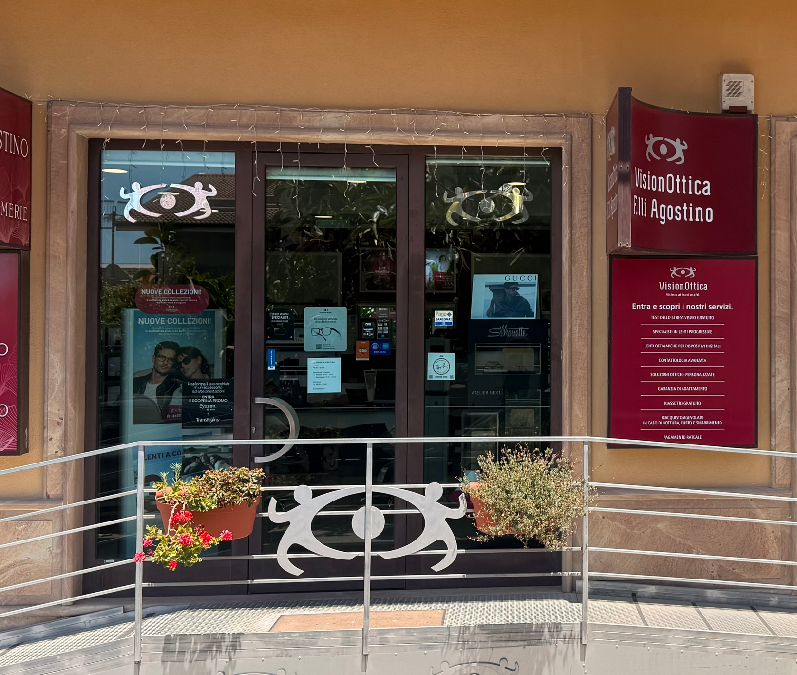Ingresso del negozio VisionOttica F.lli Agostino di Gioiosa Ionica su strada con porta a vetri scuri, logo VisionOttica sul vetro, insegna rossa laterale con lista servizi e piante decorative