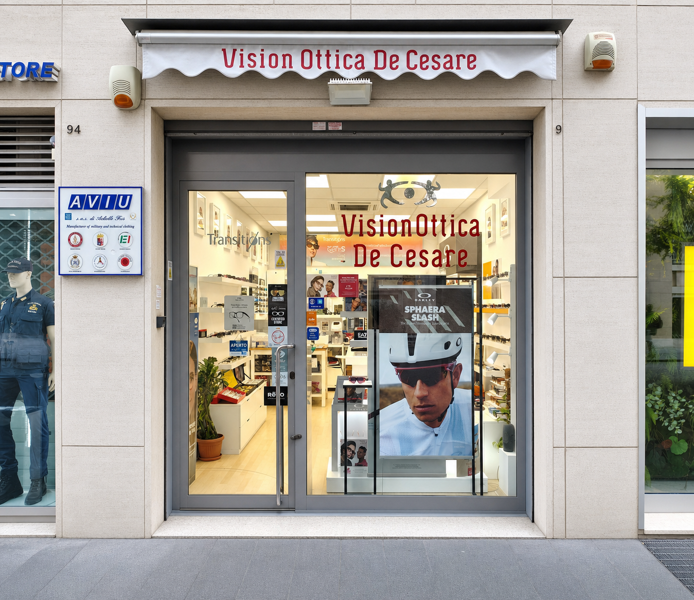 Facciata del negozio VisionOttica De Cesare di Napoli su strada con tenda a onde bianca con scritta bordeaux, porta a vetri con interno illuminato, espositori e poster brand Sphaera