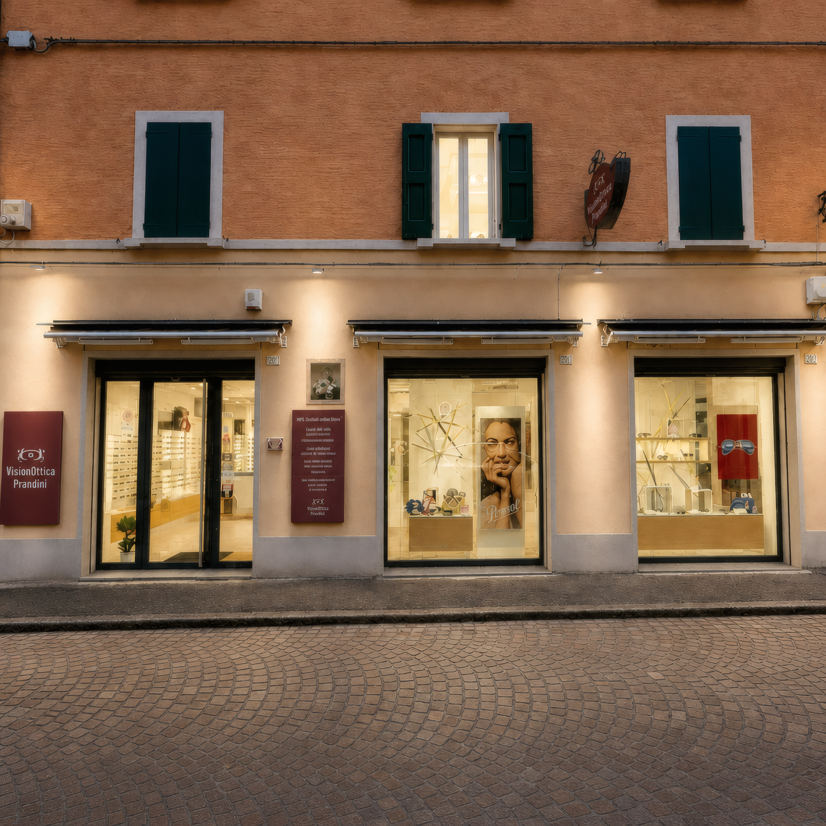 Facciata del negozio VisionOttica Prandini di Castel San Pietro Terme in centro storico su piazza lastricata di sera con edificio in mattoni, due vetrine illuminate e insegna piccola bordeaux