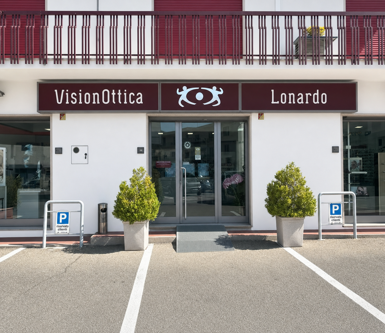 Facciata del negozio VisionOttica Lonardo di Benevento su strada con ampia insegna bordeaux bianca, porta automatica a vetri centrale, due alberelli in vaso e segnaletica parcheggio riservato clienti