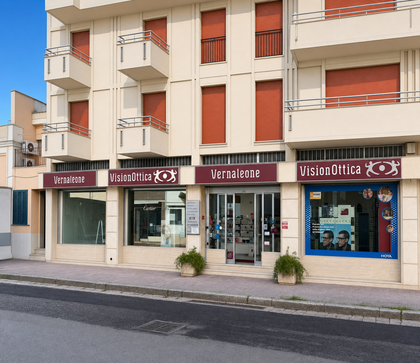 Facciata del negozio VisionOttica Vernaleone di Copertino su strada con doppia insegna bordeaux estesa su edificio residenziale, ampia vetrina con espositori e pannello Hoya laterale