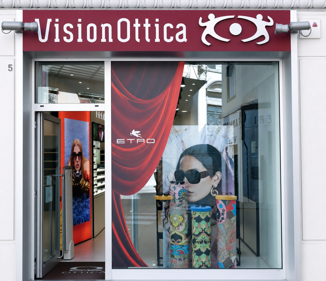 Vetrina del negozio VisionOttica Vicenza in centro su strada con insegna bordeaux, grande allestimento con tenda rossa, poster donna con occhiali da sole brand Etro e colonne decorative