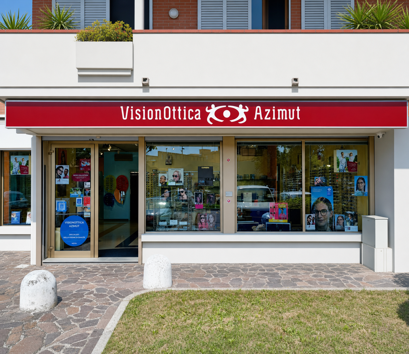 Facciata del negozio VisionOttica Azimut di Empoli su strada con tenda rossa bordeaux con insegna bianca, ampia vetrina con espositori e poster promozionali e area verde davanti